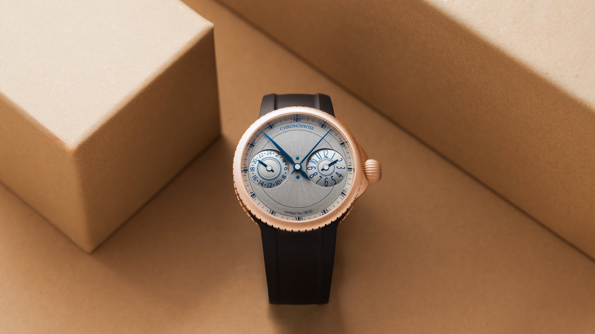 Chronoswiss Pulse GMT Frosted Guilloche Gold standing between two sand-coloured blocks, frosted dial and black rubber strap