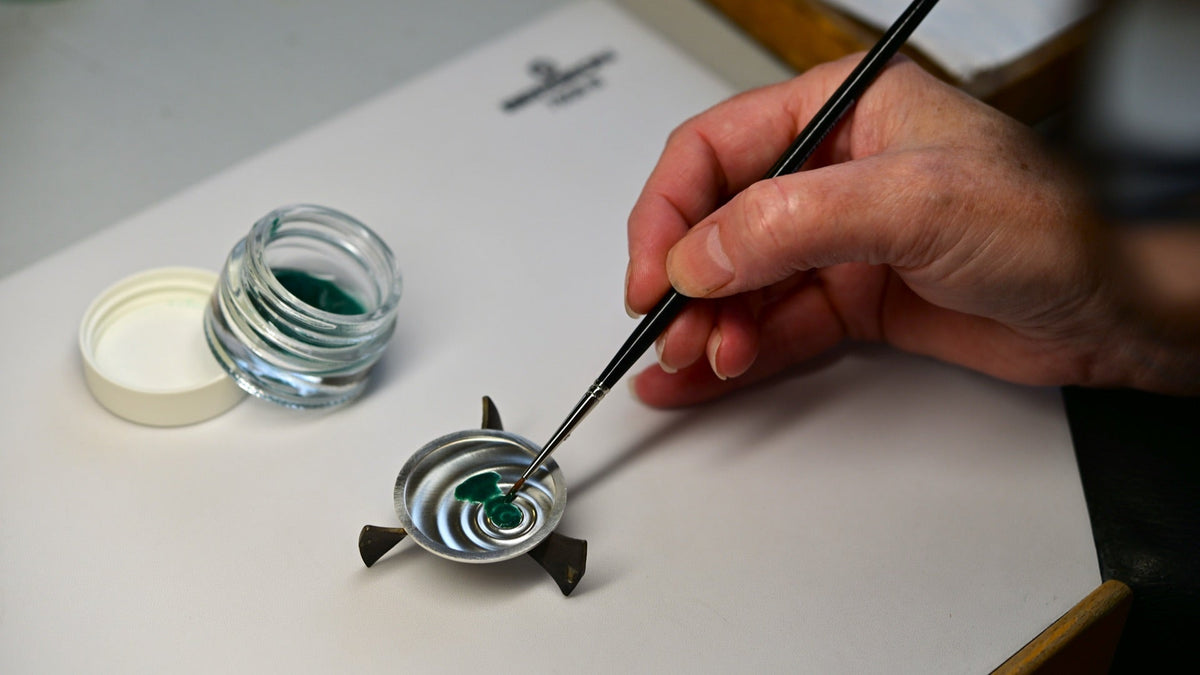 An artisan at Donzé Cadrans applies translucent green enamel to a Czapek Promenade Goutte de Rosée dial blank with a fine brush